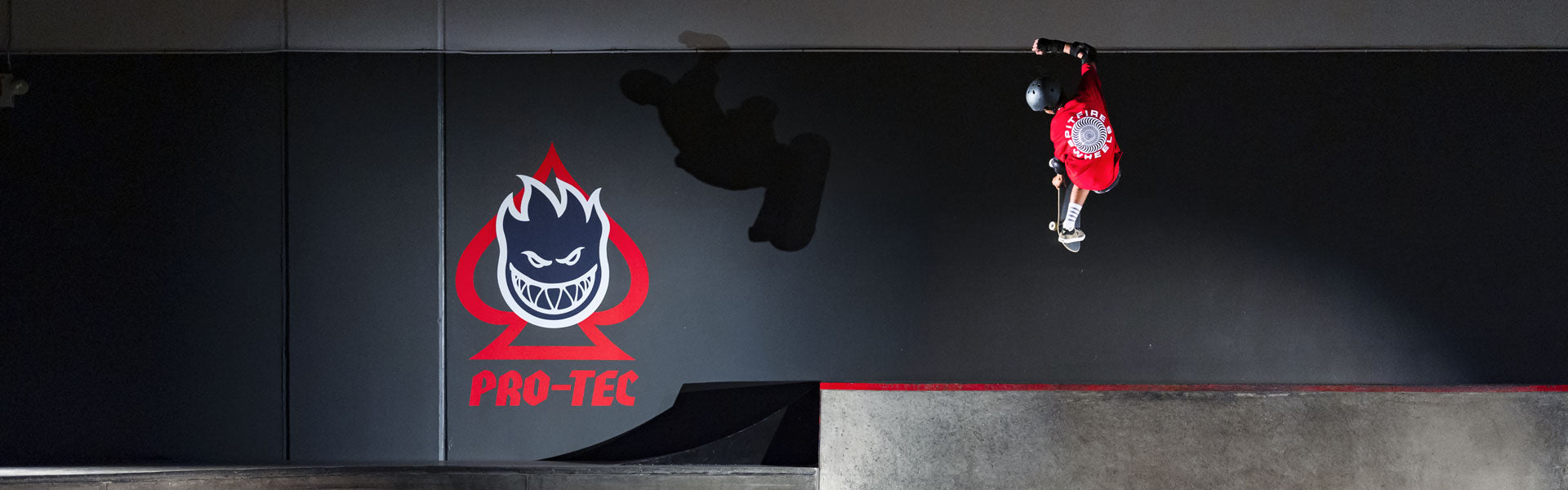 Skateboarder mid trick at a skate park with the Pro-Tec x Spitfire logo in the background.